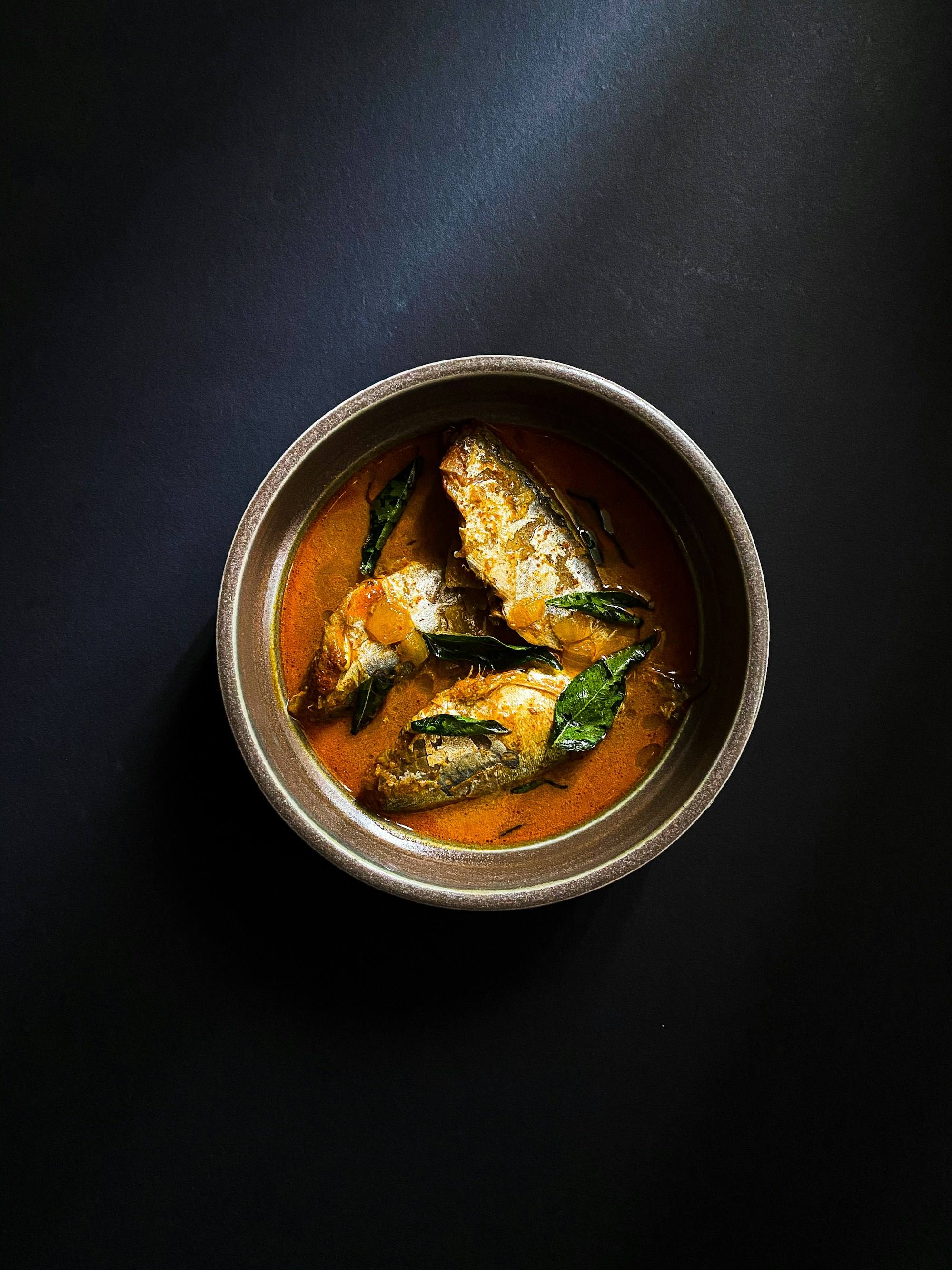 A red fish curry in a steel bowl, lit from above against a dark ground